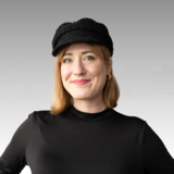 Portrait of a woman wearing a black beanie, gold earrings, and a black top against a grey background
