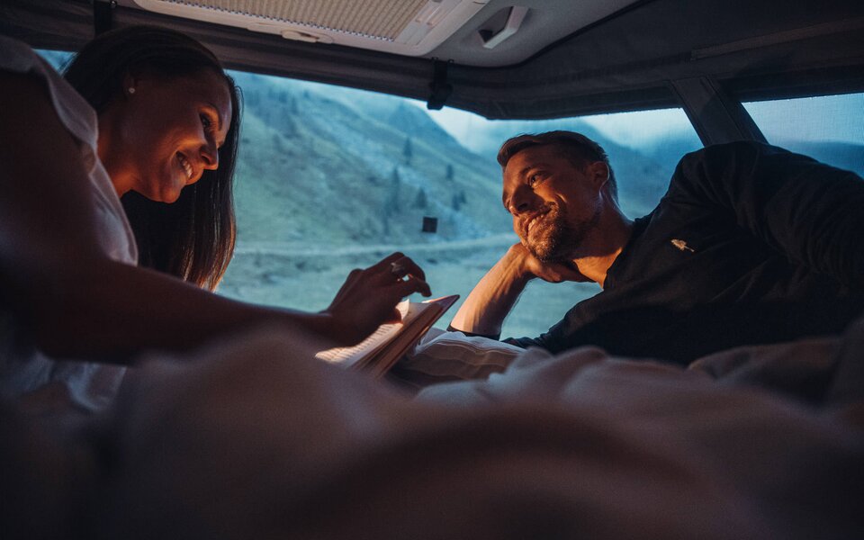 A couple lies in the bed of a Carado motorhome, gazing out at the mountain landscape at dusk