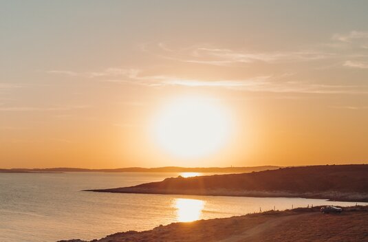 Sunset over a peaceful coastal bay, with light reflected on the water and a small vehicle by the shore
