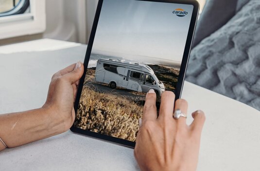 Hands holding a tablet showing a side view of a Carado motorhome on a coastal road