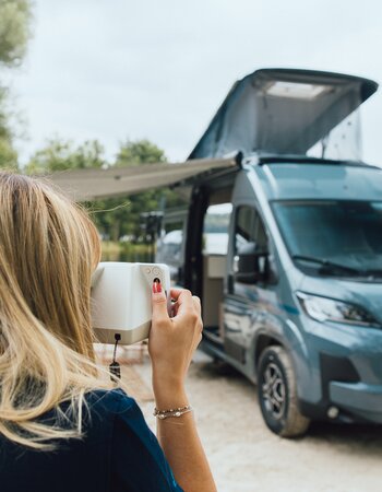 Woman photographing a Carado motorhome with pop-up roof at a lakeside pitch