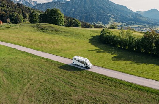Carado motorhome on a country road through green meadows with a mountain backdrop
