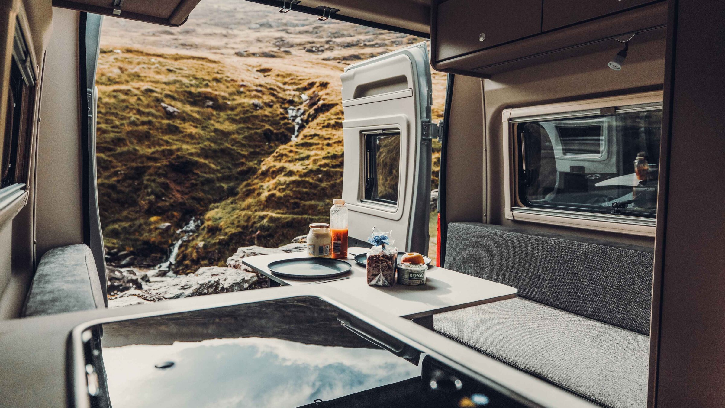 Interior of a Carado motorhome with a seating area, table and rear door open to rolling hills