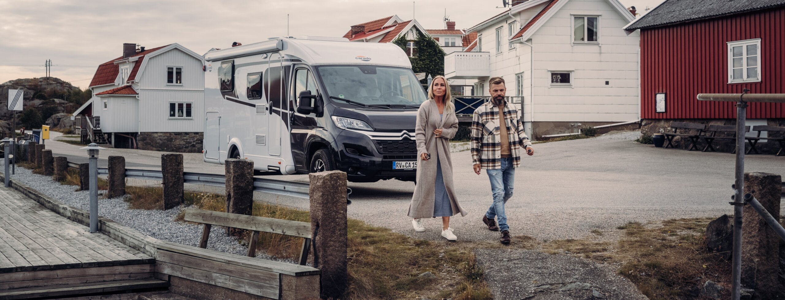 A couple walks in front of a parked Carado motorhome between houses on a coastal road