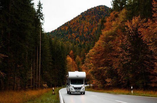 Carado motorhome on a country road through an autumn forest, with a wooded mountain in the background