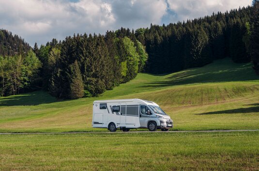 Carado motorhome in side view on a road, with green meadows and a wooded hillside in the background