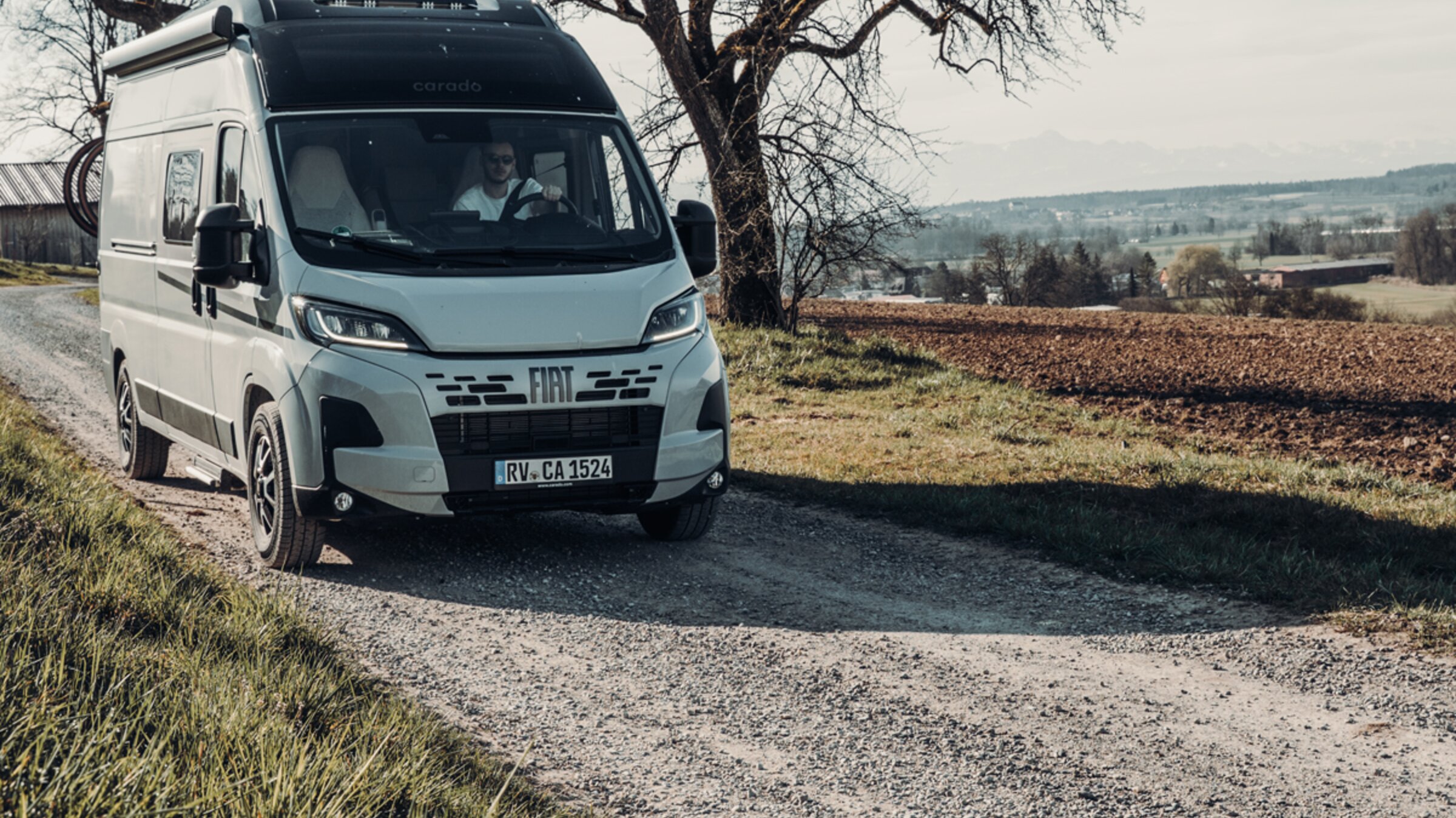 Carado Wohnmobil auf Schotterweg vor kahlen Bäumen und Feldern, Frontansicht in ländlicher Landschaft