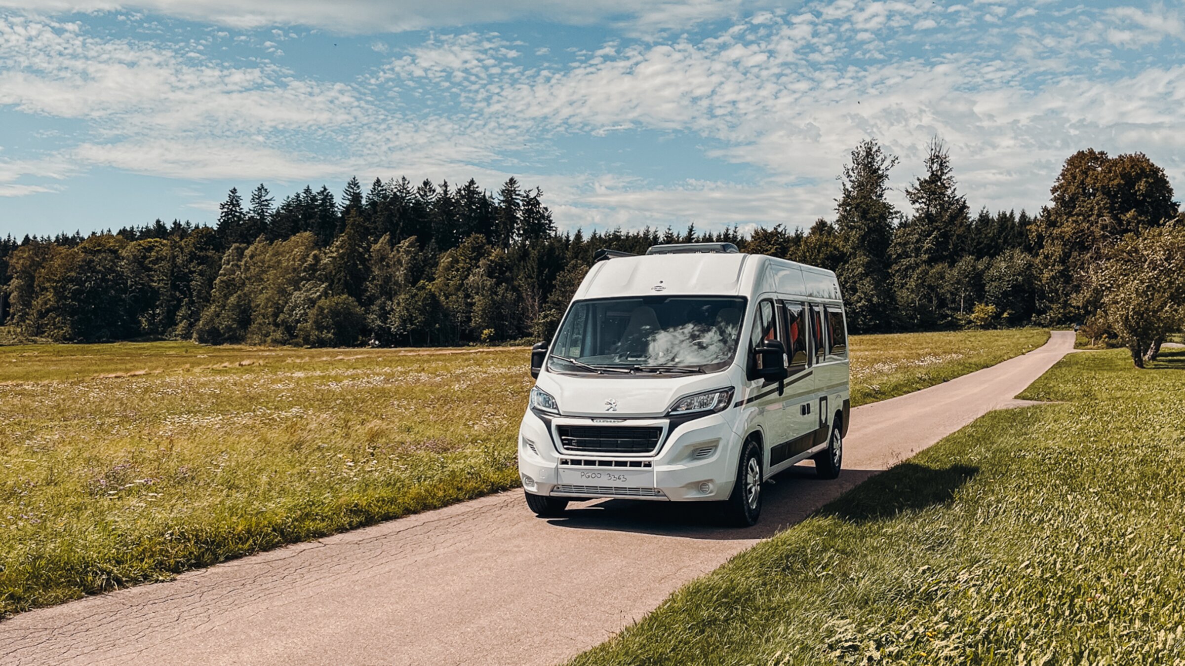 Carado Wohnmobil auf schmaler Landstraße zwischen Wiesen, Frontansicht unter blauem Himmel mit Wolken