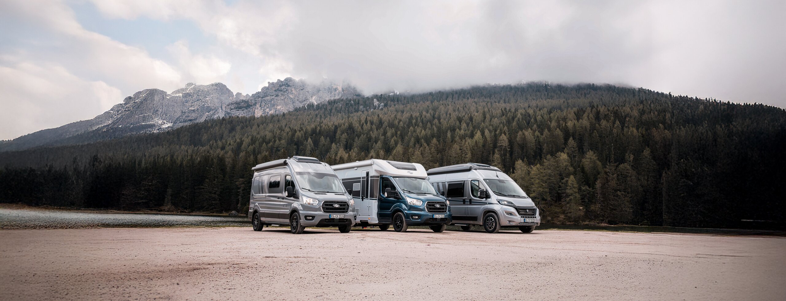 Three Carado motorhomes and camper vans on an open site in front of forest, lake and mountains