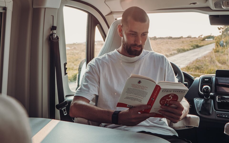 A man is reading a book in the cab of a Carado motorhome, looking through the windscreen onto a country road