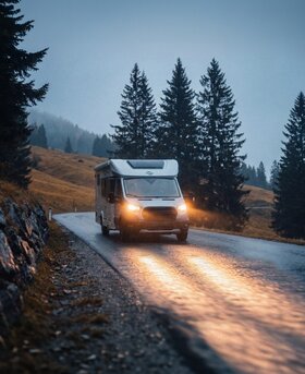 Carado motorhome with the lights on, driving along a wet country road at dusk, surrounded by fir trees and rolling hills