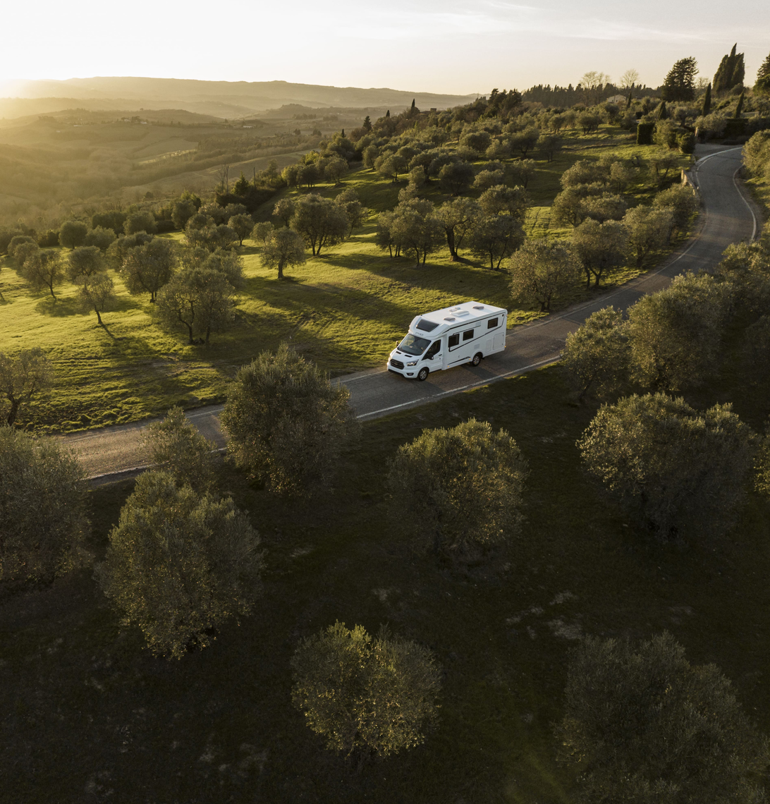 A semi-integrated Etrusco motorhome on a Ford base drives through a picturesque landscape with olive trees at golden sunset.