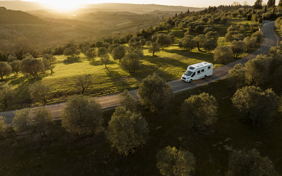 An Etrusco semi-integrated motorhome drives through a hilly landscape with olive trees and wide fields at sunset.