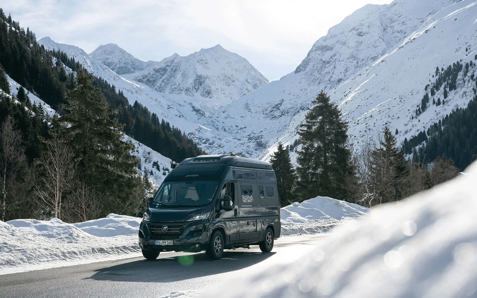 Dark HYMER camper van driving through a wooded, wintry landscape with snow-covered mountains in the background.	