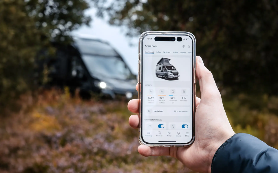 A hand holding a mobile phone displaying the content of an app for controlling a camper van. In the blurred background, a camper van can be seen in a wooded setting.	