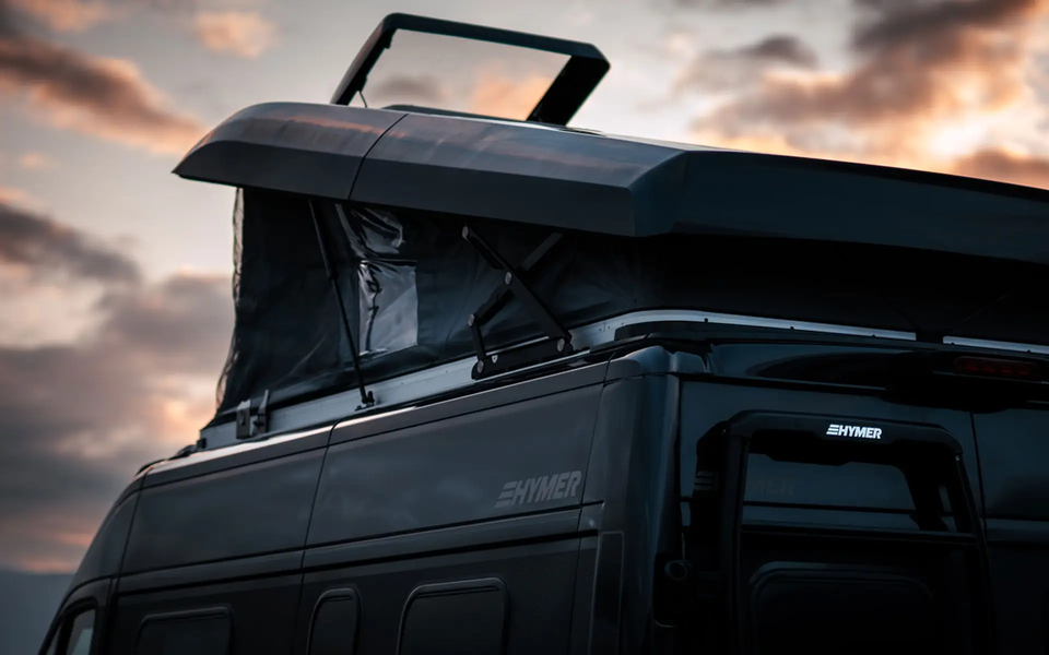 Detailed view of a black, raised pop-top roof on a black HYMER camper van against a dusky evening sky. The pop-top is seen from the rear side; you can see the shock absorber and the scissor mechanism holding the roof up.	