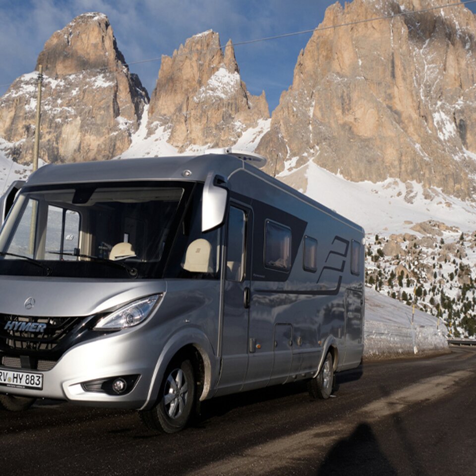 A silver HYMER B-Class MasterLine I is parked on a paved road in sunny weather, with mountains and snow in the background. 
