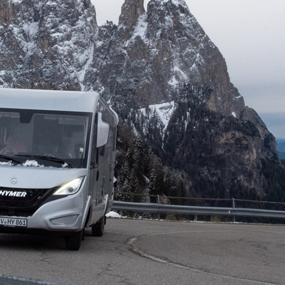 A grey HYMER B-Class ModernComfort I, a Mercedes RV, stands on a winding mountain road. There is snow on the mountains behind it.