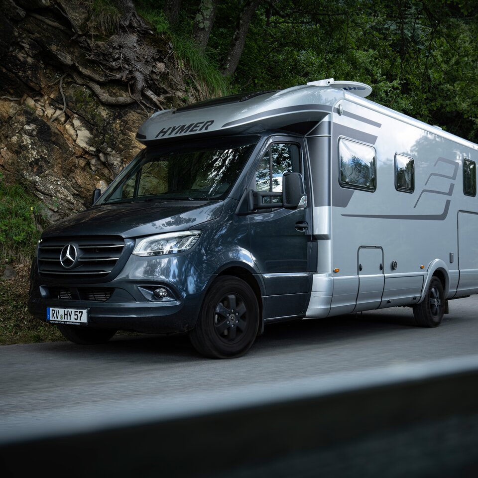The HYMER B-Class MasterLine T with a dark cab and grey body is parked on a road with a rocky embankment and green trees at the edge.