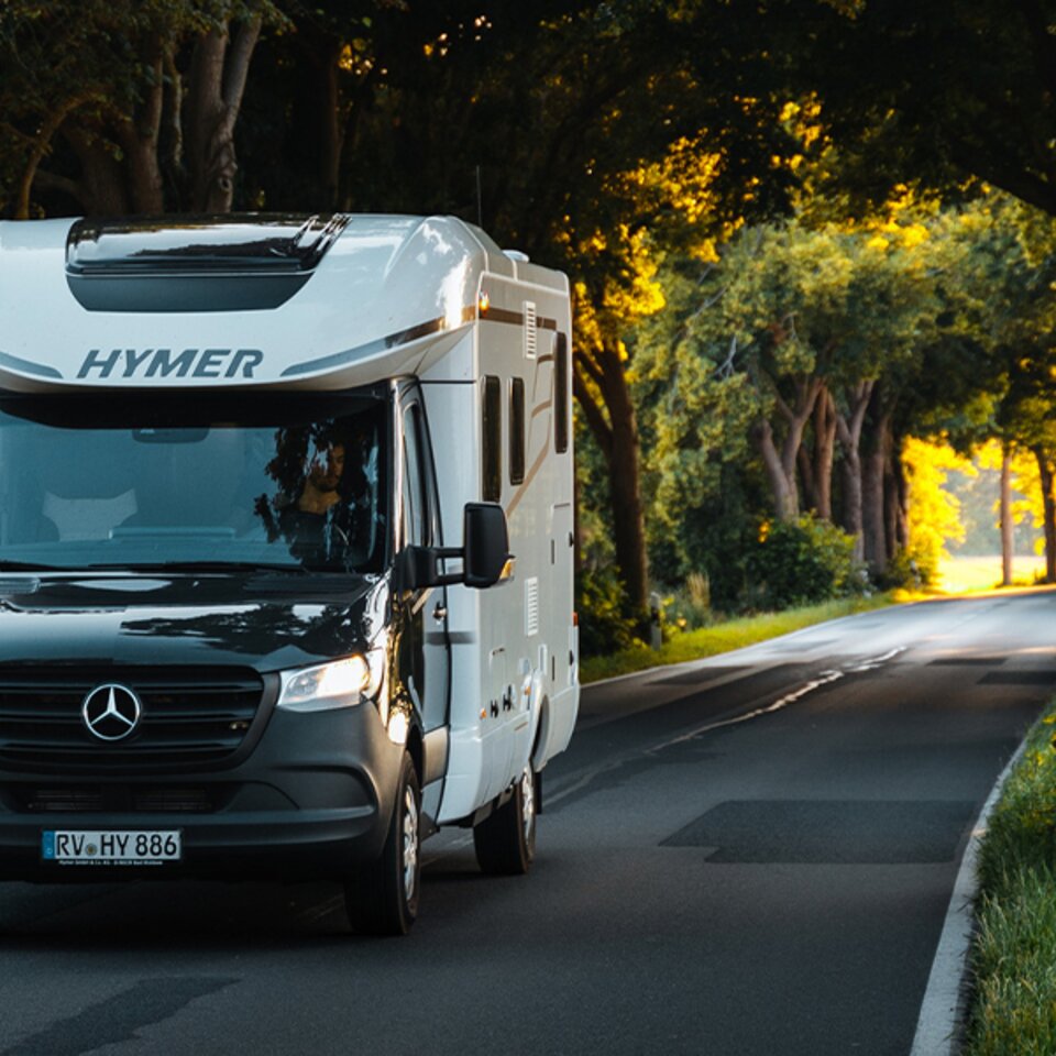 A black and white HYMER B-Class ModernComfort T with bright headlights drives through an avenue of green-leafed trees in sunny weather.