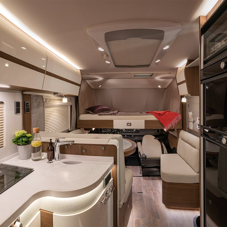 View of the Mercedes B-Class MasterLine I motorhome with wooden decor, illuminated kitchen, bench seat and bed with red blanket and purple cushion. 