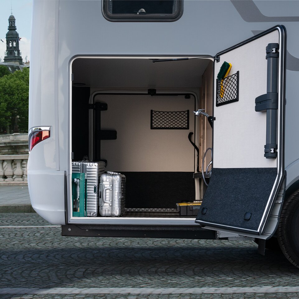 One door of the garage of the HYMER B-Class MasterLine T is open, with silver suitcases inside. The Mercedes motorhome is parked on grey paving.