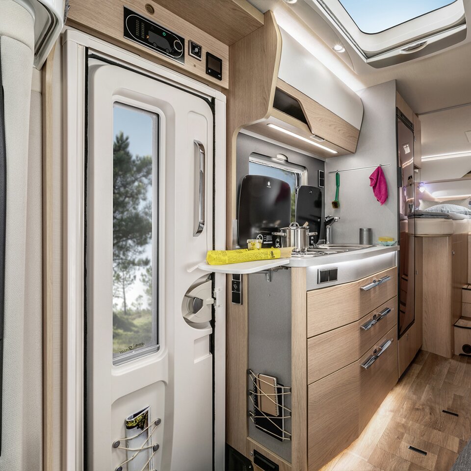 View of the cooking area of the HYMER B-Class Modern Comfort T in light-coloured wood, next to the white door with trees visible through the window.