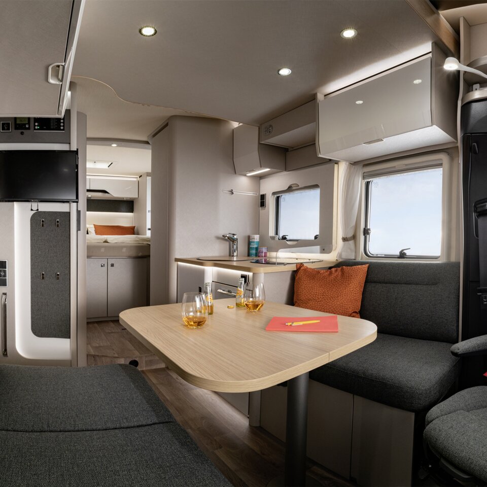 On the grey table at the grey upholstered seating area in the HYMER Tramp S are two glasses with drinks, next to them two bottles and a red notebook. 
