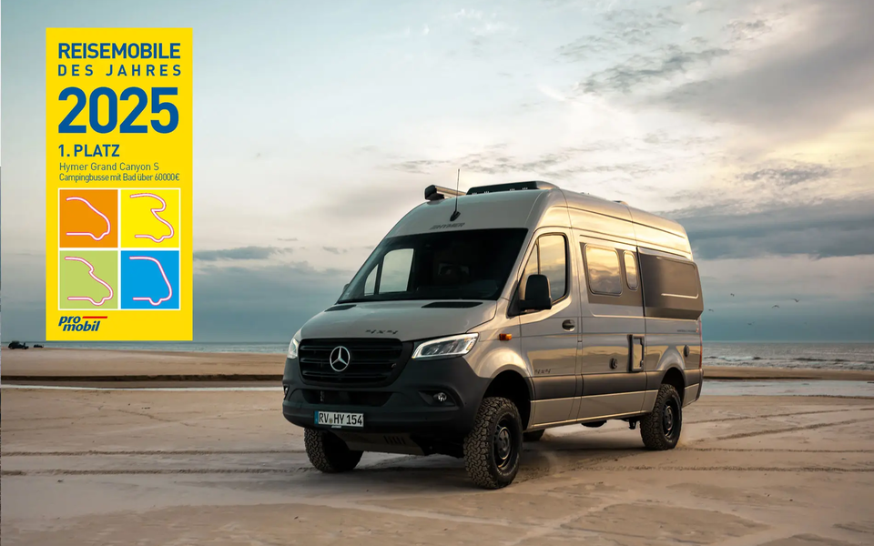 View from the front angle of a light grey HYMER 4x4 campervan parked on a sandy beach. In the background, the sea and a slightly cloudy sky can be seen. Award logo "Motorhome of the Year 2025, 1st Place Hymer Grand Canyon S" is placed on the image.	