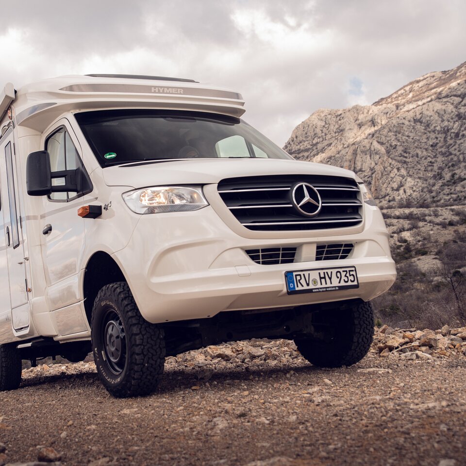 The white Mercedes HYMER ML-T all-wheel motorhome is parked on an ascending stone road with rocky hills and a slightly cloudy sky in the background.