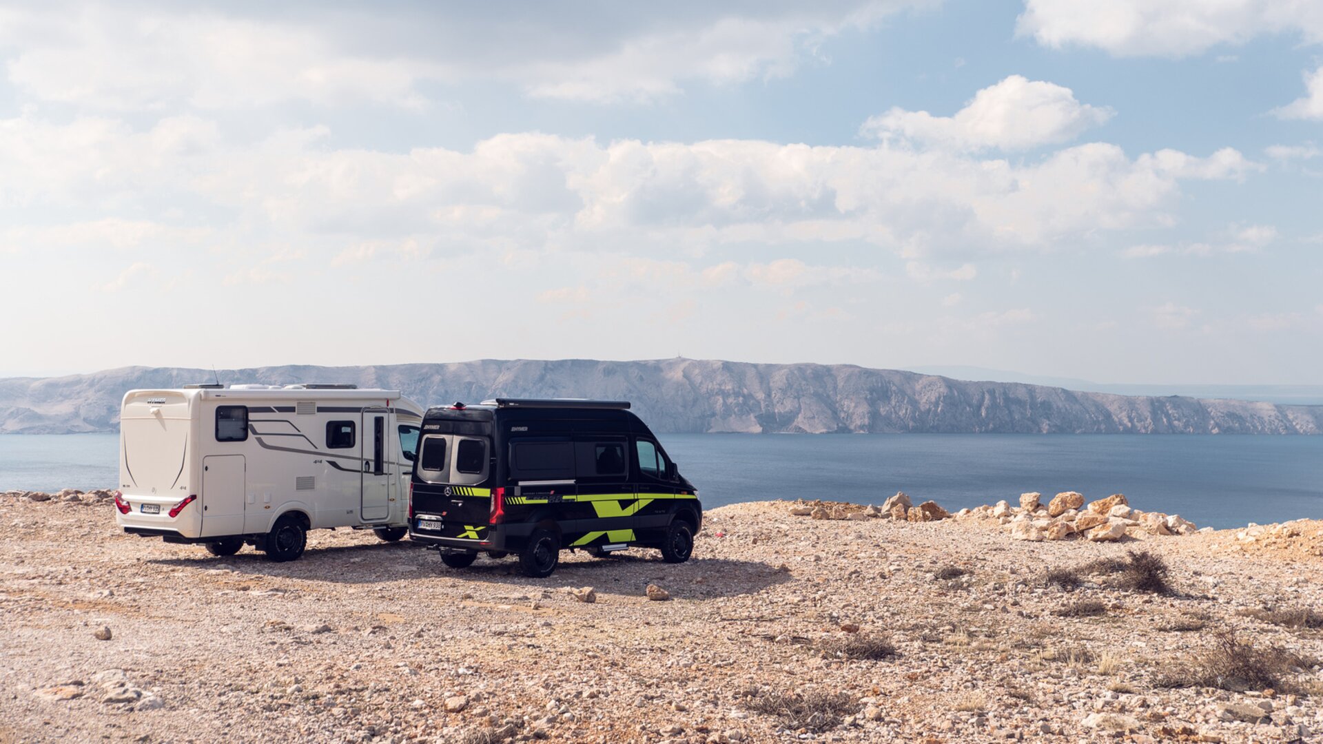 Two off-road motorhomes in white and black and green are parked on a sunlit plain above the sea, with cliffs in the background.