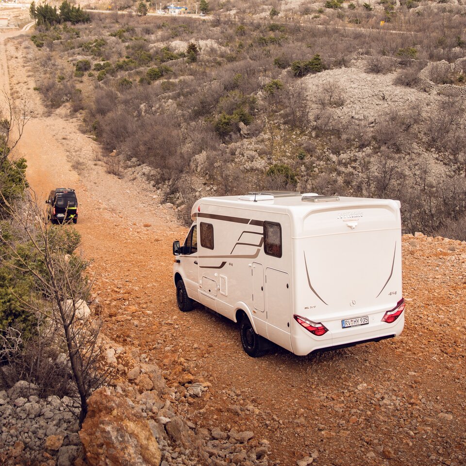 Two 4x4 off-road motorhomes from HYMER with Mercedes drive units drive on a hilly road through unpaved, stony terrain with bushes. 