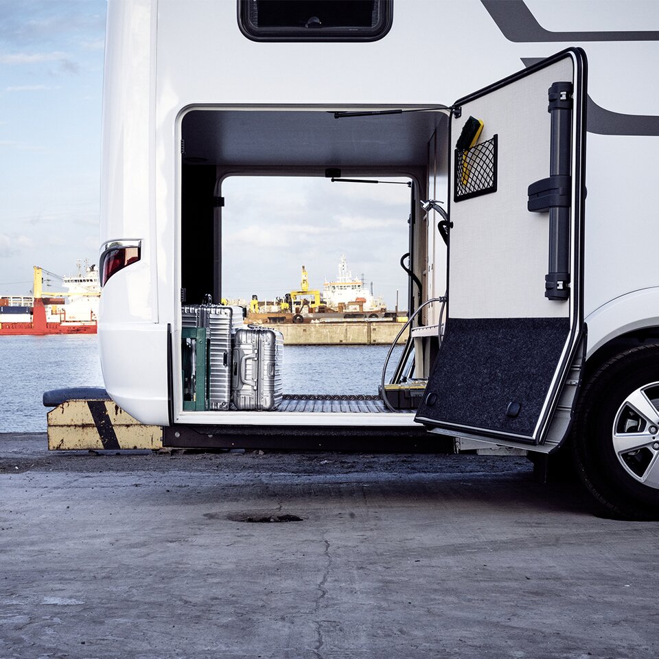There are suitcases in the open garage of the HYMER B-Class Master Line I, a motorhome with a Mercedes power unit. A harbour can be seen in the background.
