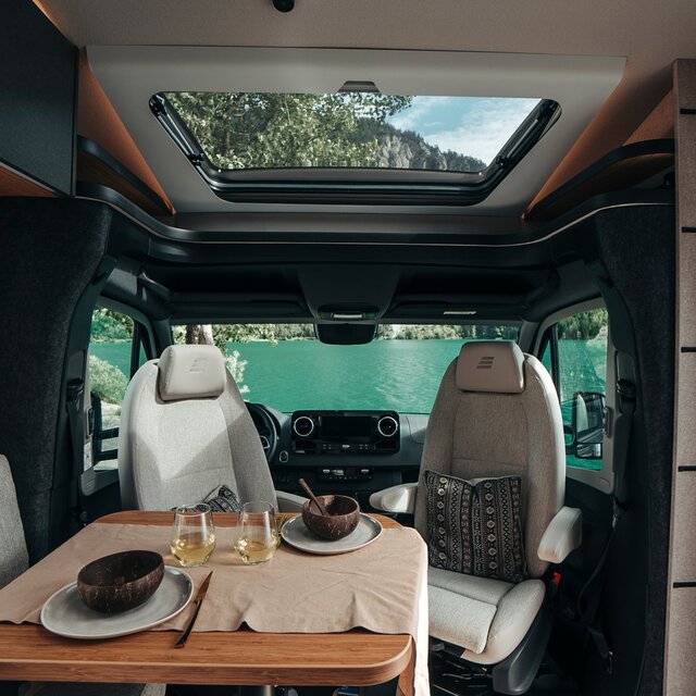 The table of the HYMER ML-T in the Native Bamboo style is set with dishes and white wine. Behind it is the driver's cabin with a view of nature.