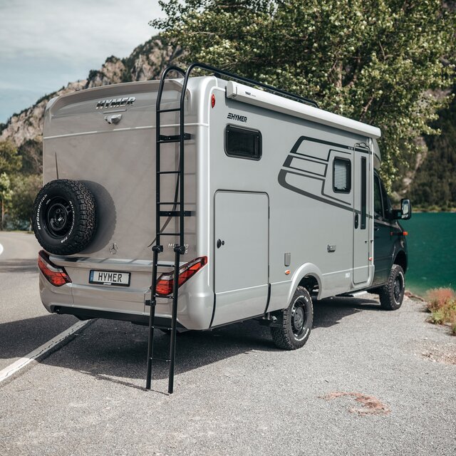 The HYMER ML-T is seen from a slight angle from behind. It is slightly turned in front of a mountain lake, and the roof ladder is extended.