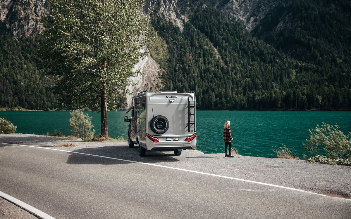 A woman stands in front of the rear of the HYMER ML-T, looking at a turquoise lake.