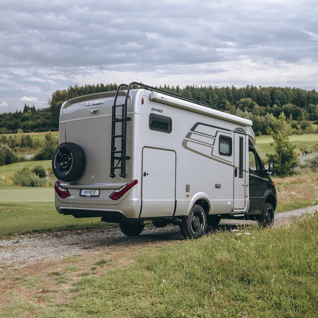The HYMER ML-T is seen from the rear, standing on a dirt road between two meadows.