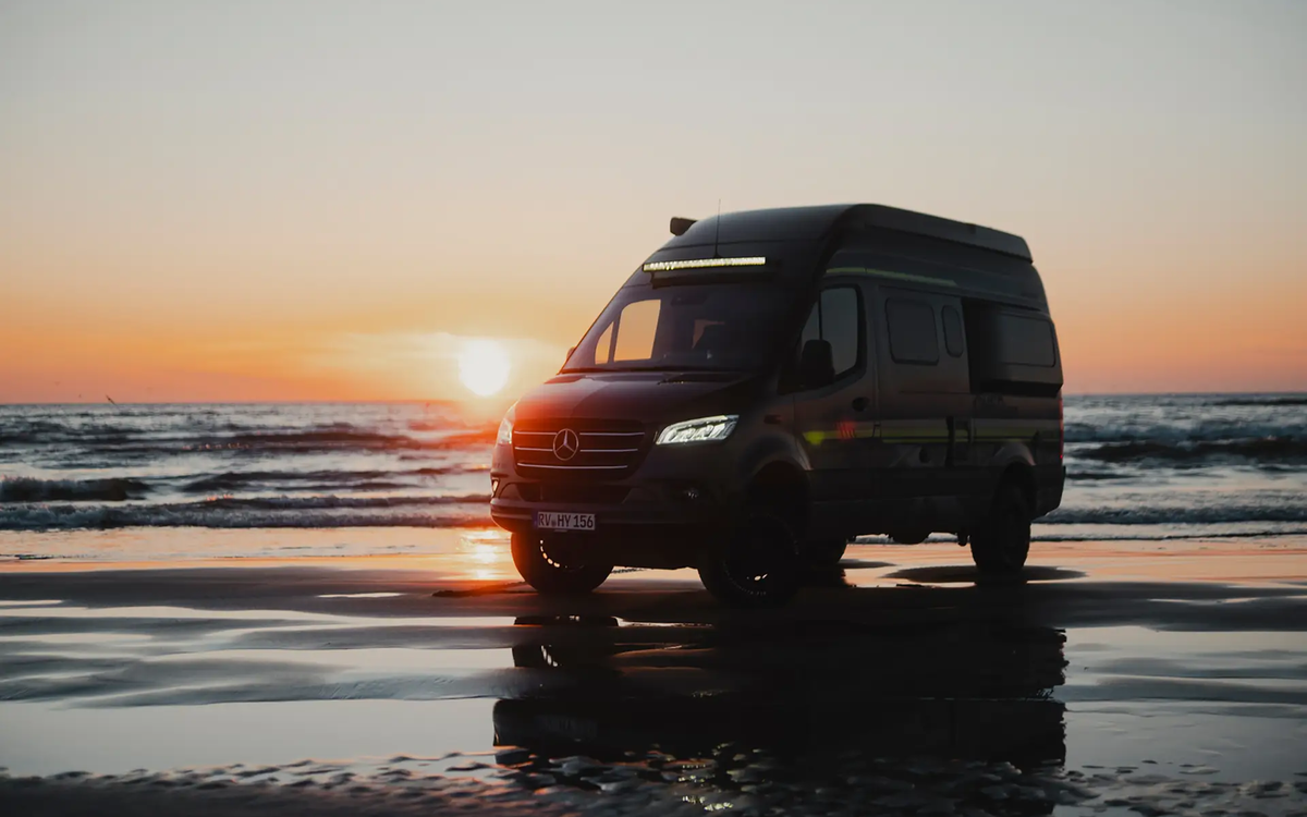 Black camper van with neon green decals stands sideways at an angle on a beach, allowing the front to be seen, with the sea in the background where the sun is setting