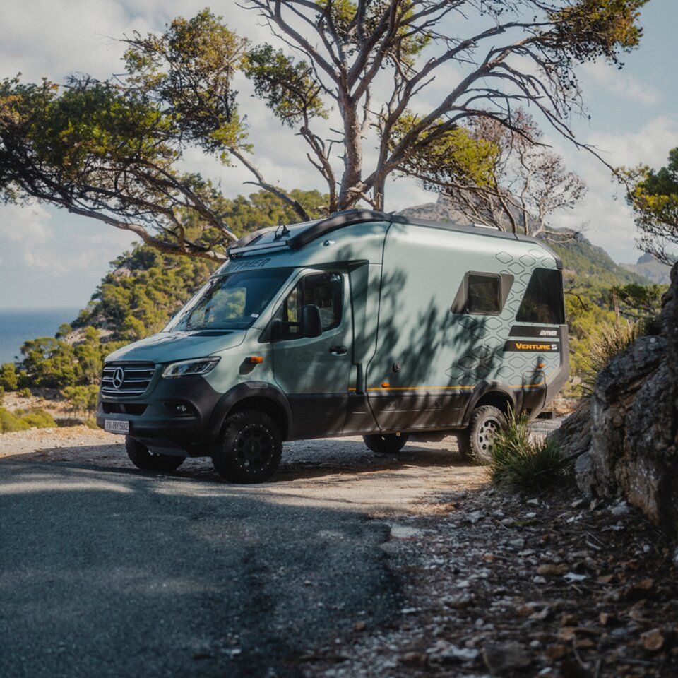 View from the front side of the driver's side of the Venture S, surrounded by trees and mountains, with the sea in the background.