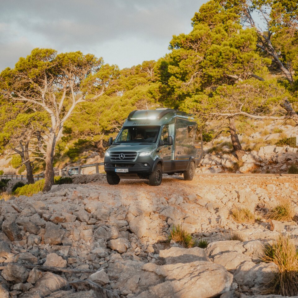 View from the front of the Venture S, headlights on, the vehicle is standing in front of a rocky abyss, surrounded by trees.