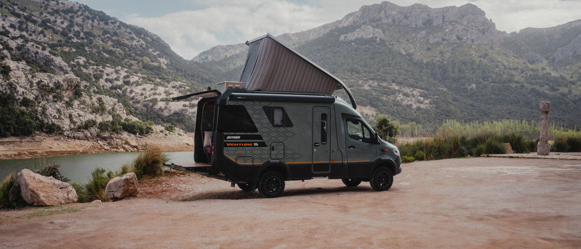 Side view of the passenger side of the Venture S, with the pop-top and Sunset Deck extended, mountains and water in the background.