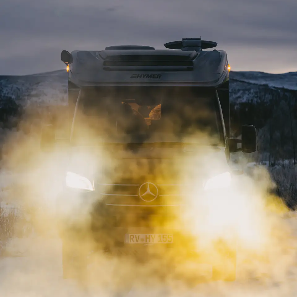 Frontudsigt over HYMER ML-T CrossOver med tændte forlygter, køretøj står i et sneklædt landskab, bjerge med sne i baggrunden