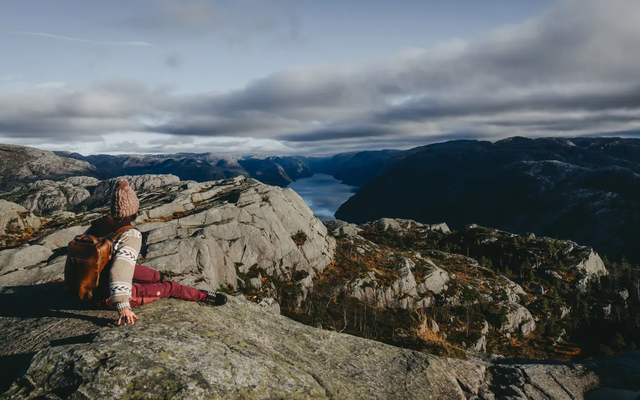 Udsigt bagfra over en vandrer, der sidder på en klippe, bjerglandskab og fjord i baggrunden