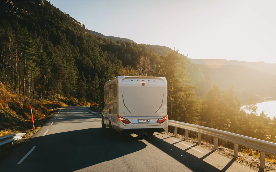 Bagudsigt af HYMER B-MC T BlackLine, køretøj kører på en landevej, skov i baggrunden til venstre, sø i baggrunden til højre