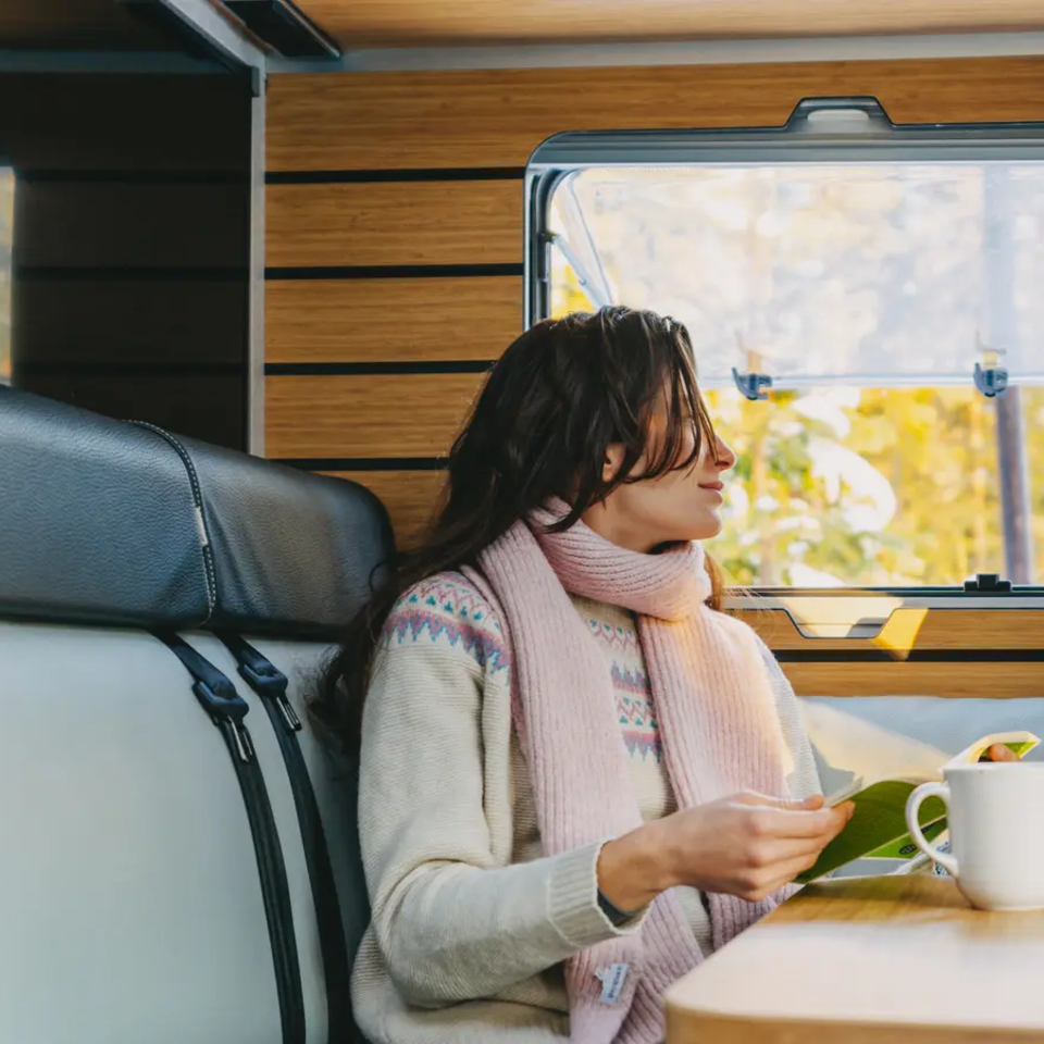 En kvinde sidder på bænkensædet i en autocamper og kigger ud af vinduet med en bog i hånden