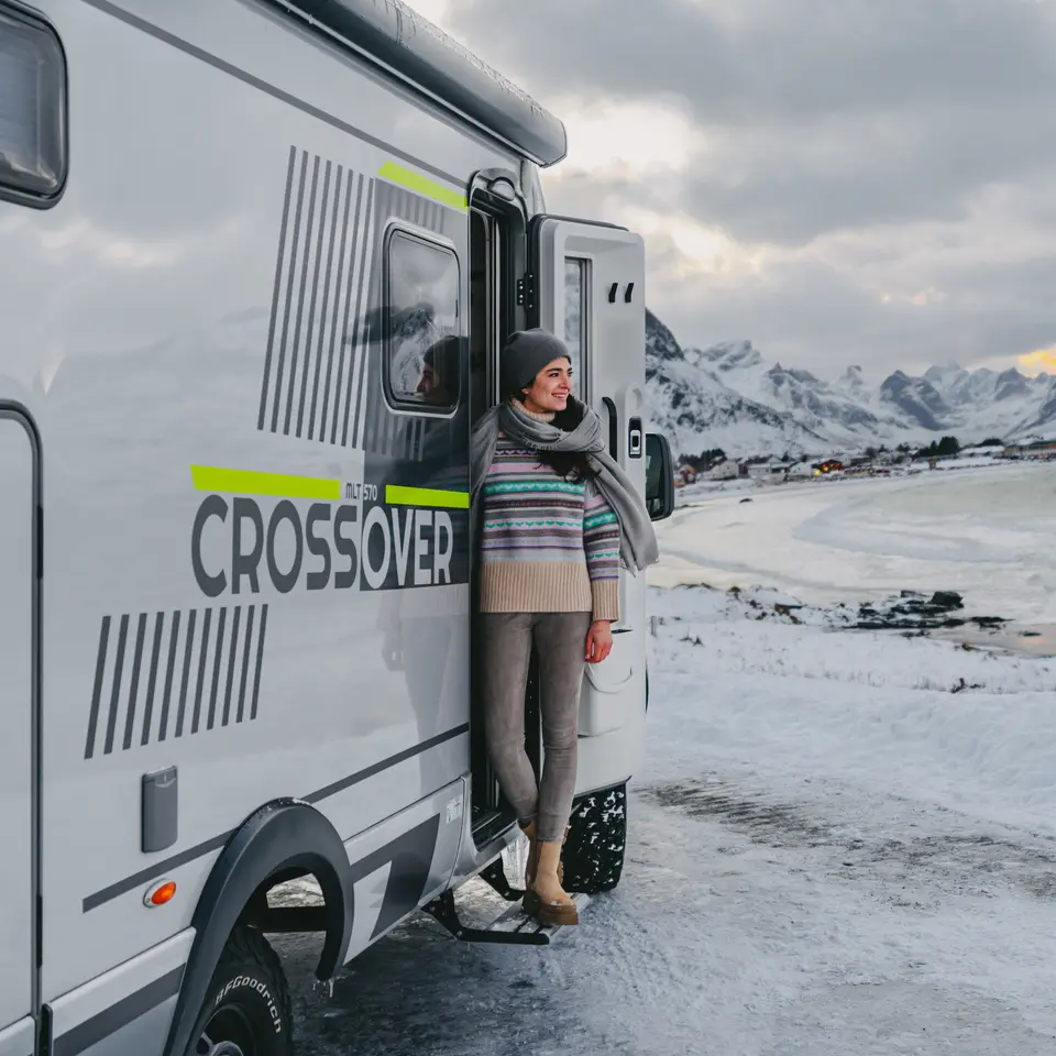 Kvinde står ved indgangsdøren til HYMER ML-T 570 CrossOver, en lille landsby ved en bugt i baggrunden