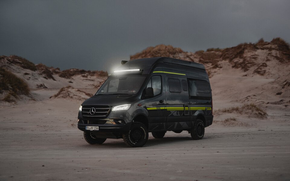HYMER off-road camper stands at dusk with headlights and light bar on a sandy beach with sand dunes in the background.