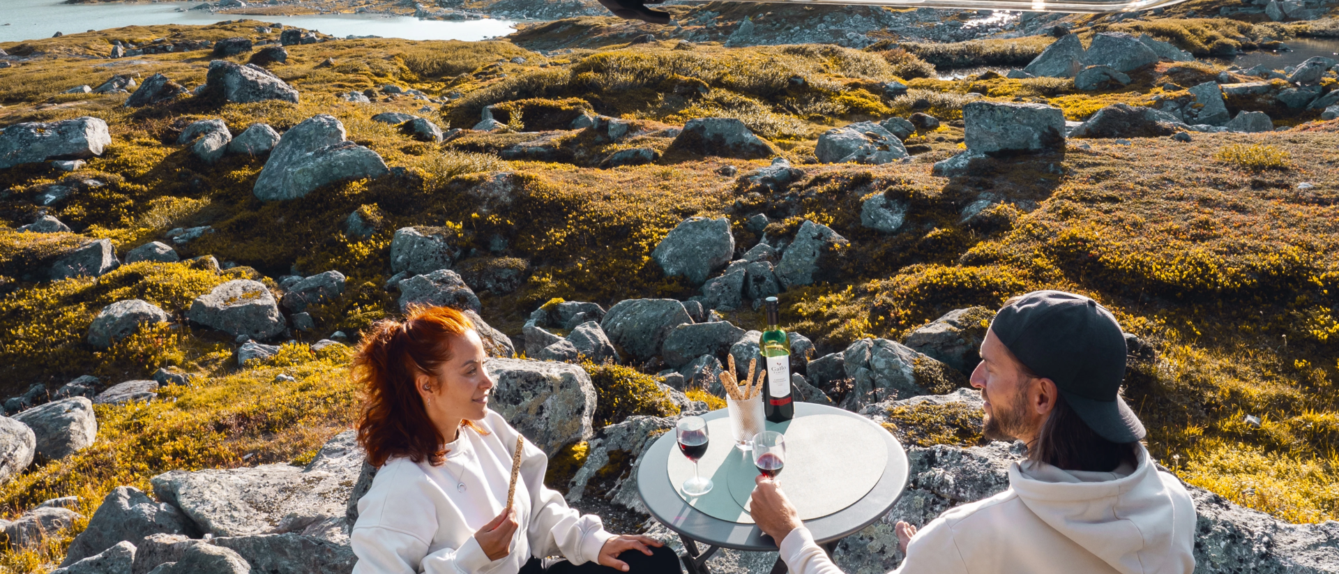 Céline et Eric assis sur des chaises à l'intérieur du LMC Innovan Active, avec une table entre eux. Ils dégustent du vin et des grissini tout en profitant de la vue sur la nature norvégienne à l'extérieur.