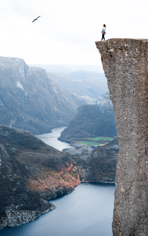 Céline debout au bord d'une falaise en Norvège, avec une vue sur les lacs et montagnes, un oiseau volant dans le ciel.