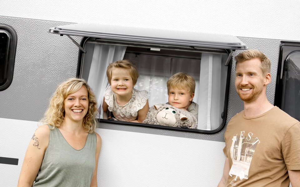 Das Bild zeigt ein Pärchen, welches vor einem LMC Sassino Wohnwagen steht, während zwei Kinder durch das geöffnete Fenster schauen.